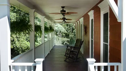 Sunny porch with white railing, wooden floor, ceiling fans, hanging plants, and rocking chairs on a house.