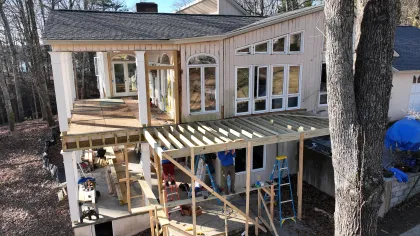 Two-story house undergoing wooden deck construction with workers, ladders, and forest surroundings on a clear day