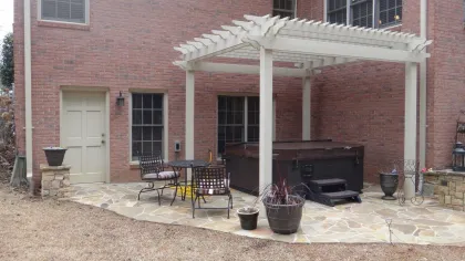 Backyard patio with stone flooring, white pergola, hot tub, metal chairs, and potted plants against a brick house.