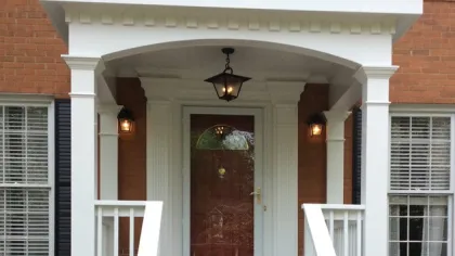 Brick house entrance with stone steps, white porch columns and railing, wooden door, and outdoor lanterns.