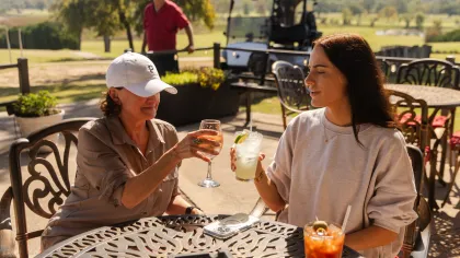 Two women clinking drinks together at an outdoor patio table on a sunny day near a golf course.