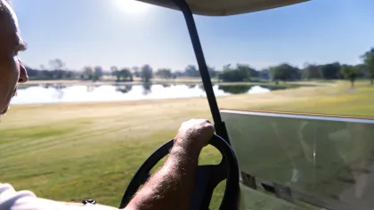 Man driving a golf cart on a sunny golf course near a water hazard with trees in the distance.