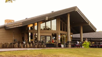 Modern lodge-style building with large windows and outdoor seating on a grassy lawn under a sloped roof.