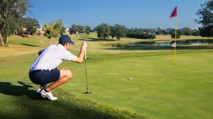 Golfer in a cap crouching and lining up a putt on a sunny golf course near the hole with a red flag.