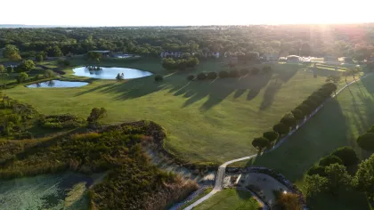Aerial view of a golf course with ponds, green fairways, trees casting long shadows at sunset.