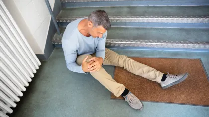 Man sitting on floor at bottom of stairs holding knee in pain next to radiator and doormat.