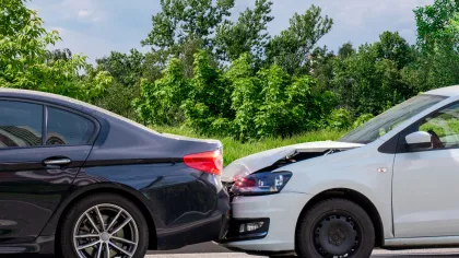 White car with front-end damage collides with black car's rear on a road with green trees and blue sky background