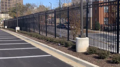 Black metal fencing alongside a parking lot, with pavement and greenery under a clear blue sky.