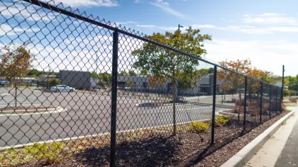 A chain-link fence borders a parking lot with trees and blue skies in the background.