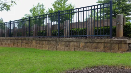 Black metal fence with vertical bars on a stone retaining wall with green grass and trees in background.