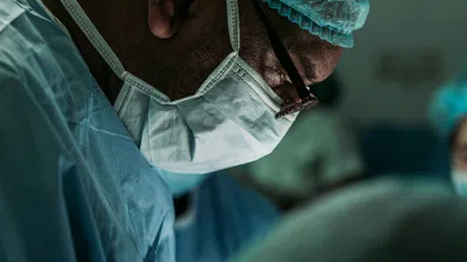 Surgeon wearing surgical mask, cap, and gown focusing during an operation in a dimly lit operating room.