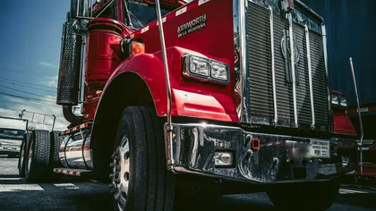 Red Kenworth semi-truck parked outdoors under a blue sky with visible chrome grille and exhaust stack.