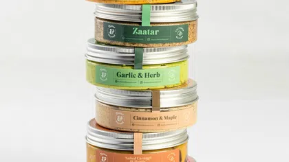 Stack of six colorful gourmet spread jars with flavors like Raspberry, Salted Caramel, Cinnamon, Garlic Herb, Zaatar, and Chilli Lime.