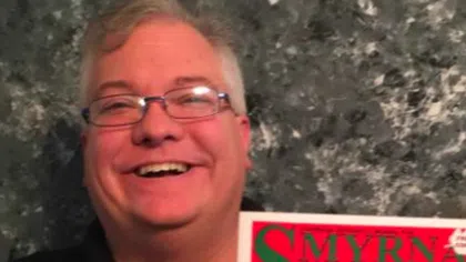 Smiling man with glasses holding a colorful Smyrna Guide publication featuring local ads and guides.