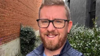 Smiling man with glasses and beard wearing a blue quarter-zip sweater standing outdoors by brick and black walls.