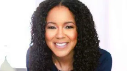 Smiling woman with curly hair in navy top leaning forward with hands clasped in bright indoor setting