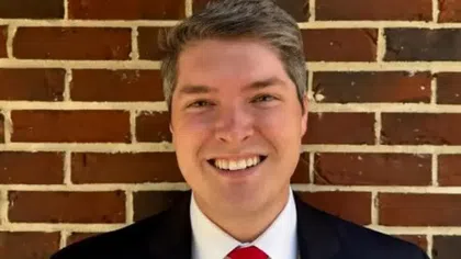 Smiling man in a black suit, white shirt, and red tie standing in front of a brick wall background