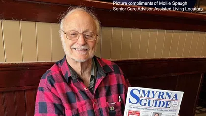 Elderly man in red plaid shirt smiling and holding a Smyrna Guide newsletter while seated on a wooden bench.