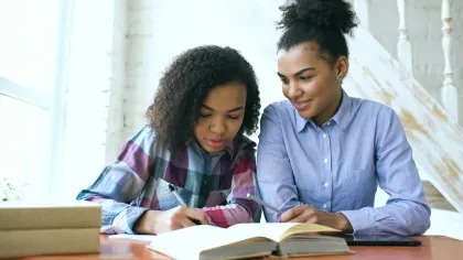 Two young women studying together at a table with open books and a tablet in a bright room