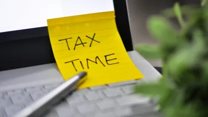 Yellow sticky note with TAX TIME written on laptop keyboard next to pen and blurred plant.