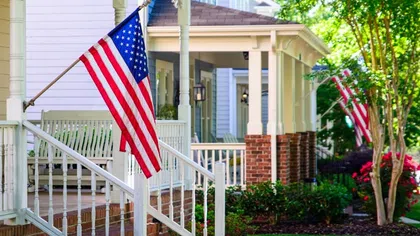American flags on porches of charming suburban homes with green lawns and blooming flowers on a sunny day