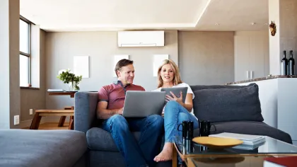Couple sitting on gray sofa in modern living room using laptop and tablet, relaxed barefoot at home.