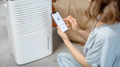 Woman controlling white dehumidifier using smartphone app while sitting on floor indoors.