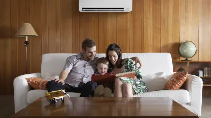 Family of three sitting on a white sofa reading a book in a cozy wooden-paneled living room with an air conditioner.