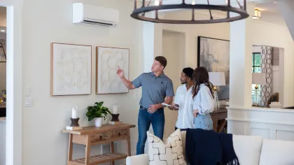 Man explains wall-mounted air conditioner to couple in modern living room with neutral decor and wooden furniture.
