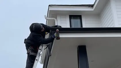 Worker on a ladder using a drill to repair the gutter of a white house with a porch and pillar.
