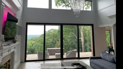 Modern living room with large glass sliding doors, outdoor patio, fireplace, chandelier, and forest views.