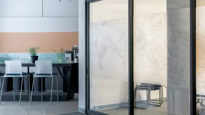 Modern office lounge with high chairs, glass partition featuring abstract map design, and coffee machine area.