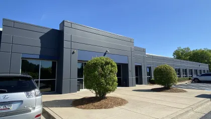 Modern gray commercial building with large windows, two trimmed bushes, and parked cars under clear blue sky.