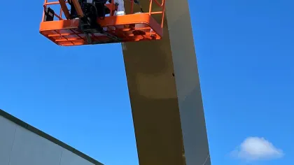 Worker in orange lift painting a tall metallic golden structure against a clear blue sky