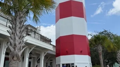 Red and white striped lighthouse near palm trees and building under a partly cloudy sky with people walking by.