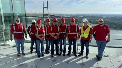 Group of construction workers in red safety vests and helmets standing on rooftop with bridge and river in background