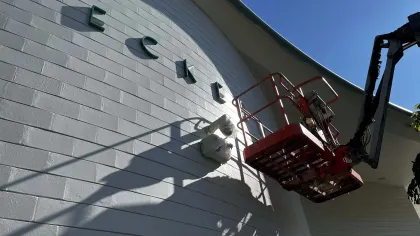 Red aerial work platform near curved white building wall with large letters mounted under clear blue sky.
