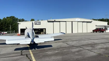 Small private airplane parked on runway in front of white FBO hangar under clear blue sky