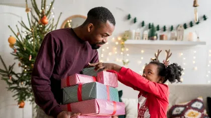 Father and daughter exchanging Christmas gifts in cozy living room decorated with tree and lights.