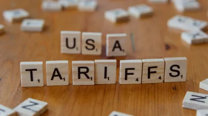 Scrabble tiles spelling USA tariffs on a wooden surface with scattered tiles around.