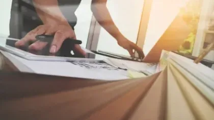 Person using a tablet and laptop with architectural blueprints and a notebook on a wooden table in sunlight.
