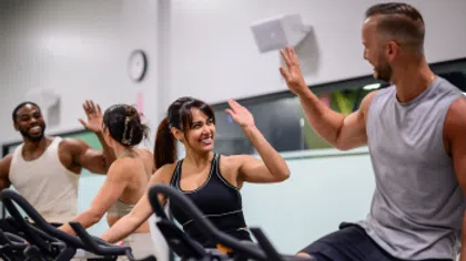 Four diverse adults on stationary bikes giving high fives in a bright gym during group cycling class