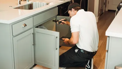 Technician inspecting plumbing under kitchen sink with flashlight in modern home kitchen.