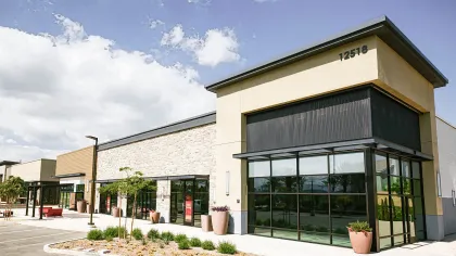 Modern commercial retail building with large glass windows, potted plants, and a clear blue sky above.