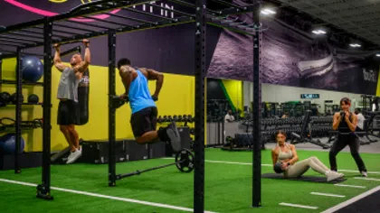 Four people exercising with weights and pull-up bars in a modern gym with green turf and purple walls.