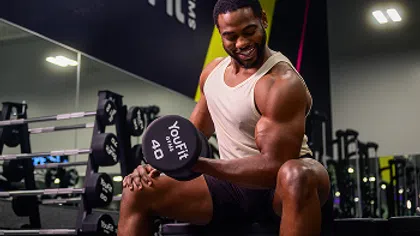 Smiling man lifting a 40-pound dumbbell while seated in a modern gym with weight racks in the background