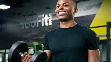 Smiling man lifting dumbbells in a modern gym with YouFit branding in the background