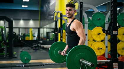 Man in black tank top lifting green barbell weights in a gym with colorful weight plates in the background.