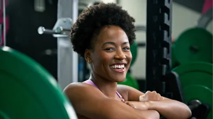 Smiling woman resting on a barbell rack in a gym surrounded by weight plates and fitness equipment.