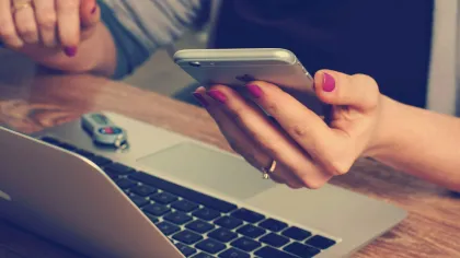 Person holding smartphone with pink nails over open laptop on wooden table, key fob beside laptop.